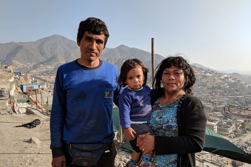 Father standing beside mother holding their son with a mountain range and city in the background.