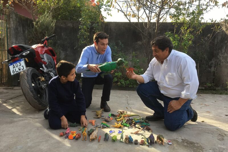 Two men and a child knelt on the ground playing with dinosaur toys