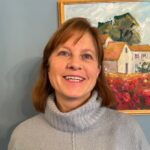 Gratitude-Suzy-Donor-Feature A White woman is smiling and standing in front of a painting of a house