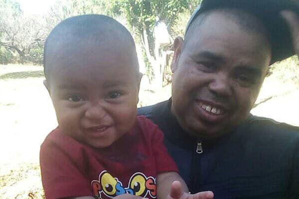 A Black baby boy and his father are standing in a green garden and smiling at the camera.