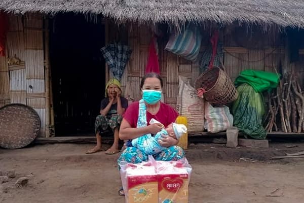 A woman wearing a mask is feeding a baby outside of a thatched hut