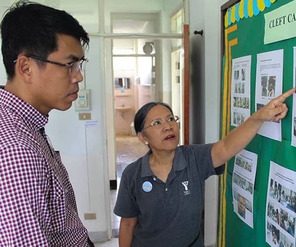 An older woman is pointing to a Cleft Care bulletin board and showing a man image of children that are pinned to the board.