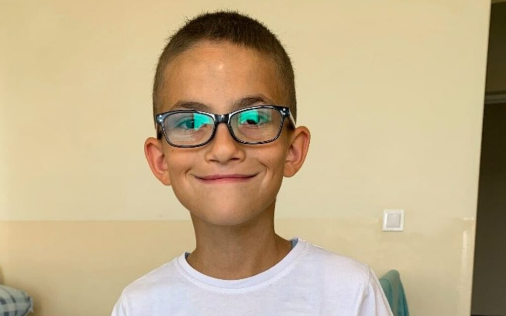 CreativeCloud 2022-05-04 14.15.29 A young boy with glasses and short hair standing against a yellow wall.